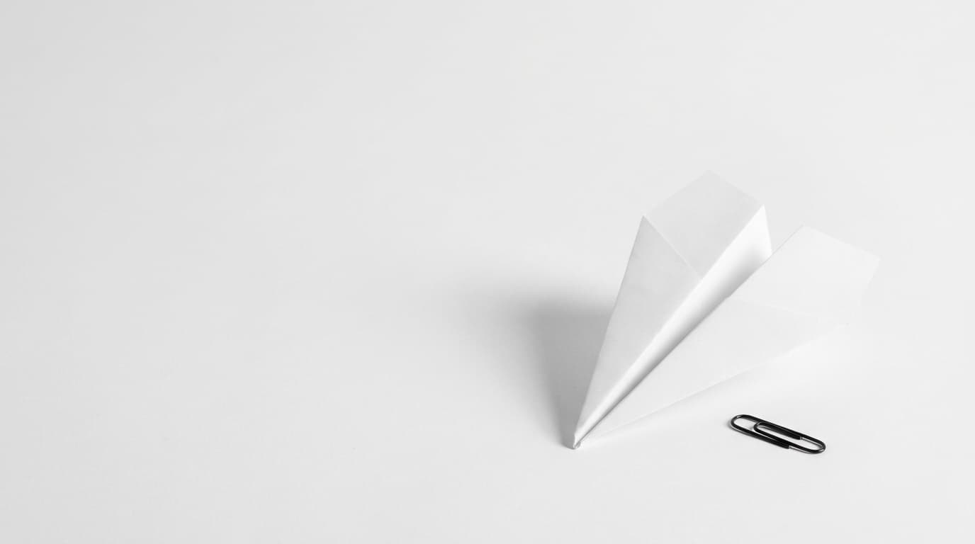 A white paper airplane on white, almost invisible, with a single black paperclip